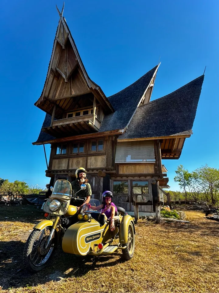 Guests on a Royal Enfield sidecar in front of a traditional wooden house with soaring roofs